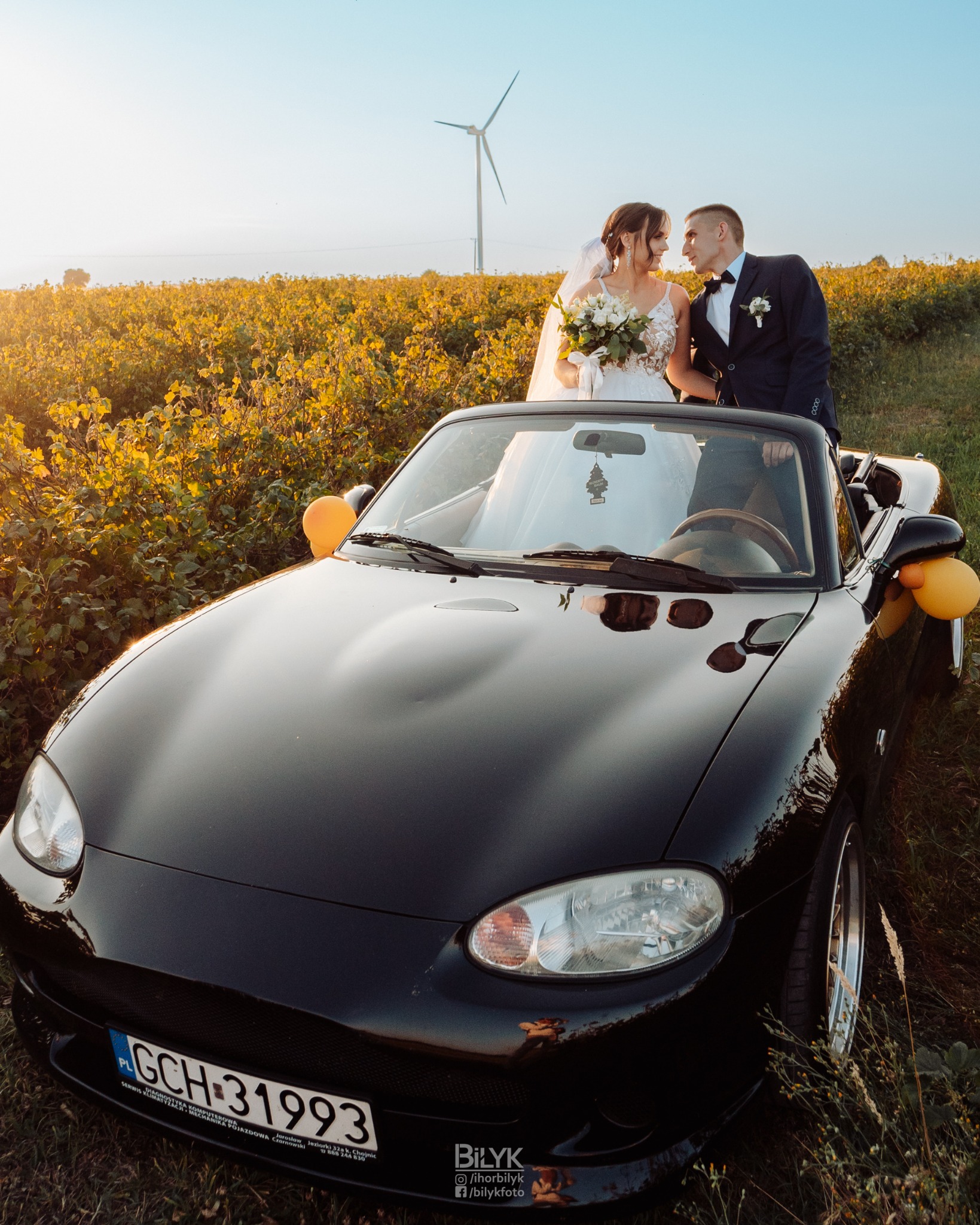 Para młoda w strojach ślubnych pozuje przy czarnym kabriolecie Mazda MX-5 z żółtymi balonami, na tle pola i wiatraka.