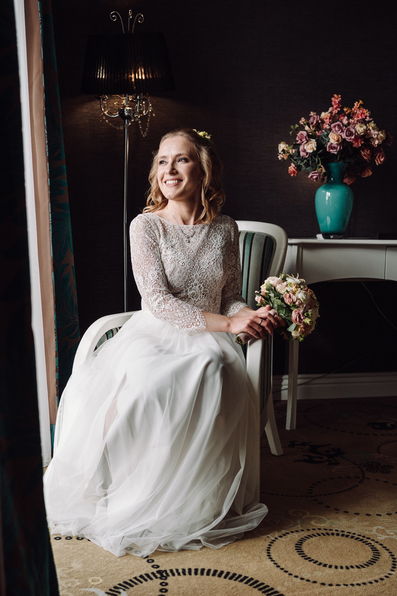 Uśmiechnięta panna młoda w koronkowej sukni siedzi z bukietem na krześle obok okna, z kwiatami w wazonie w tle. Eleganckie wnętrze, ciepłe światło.