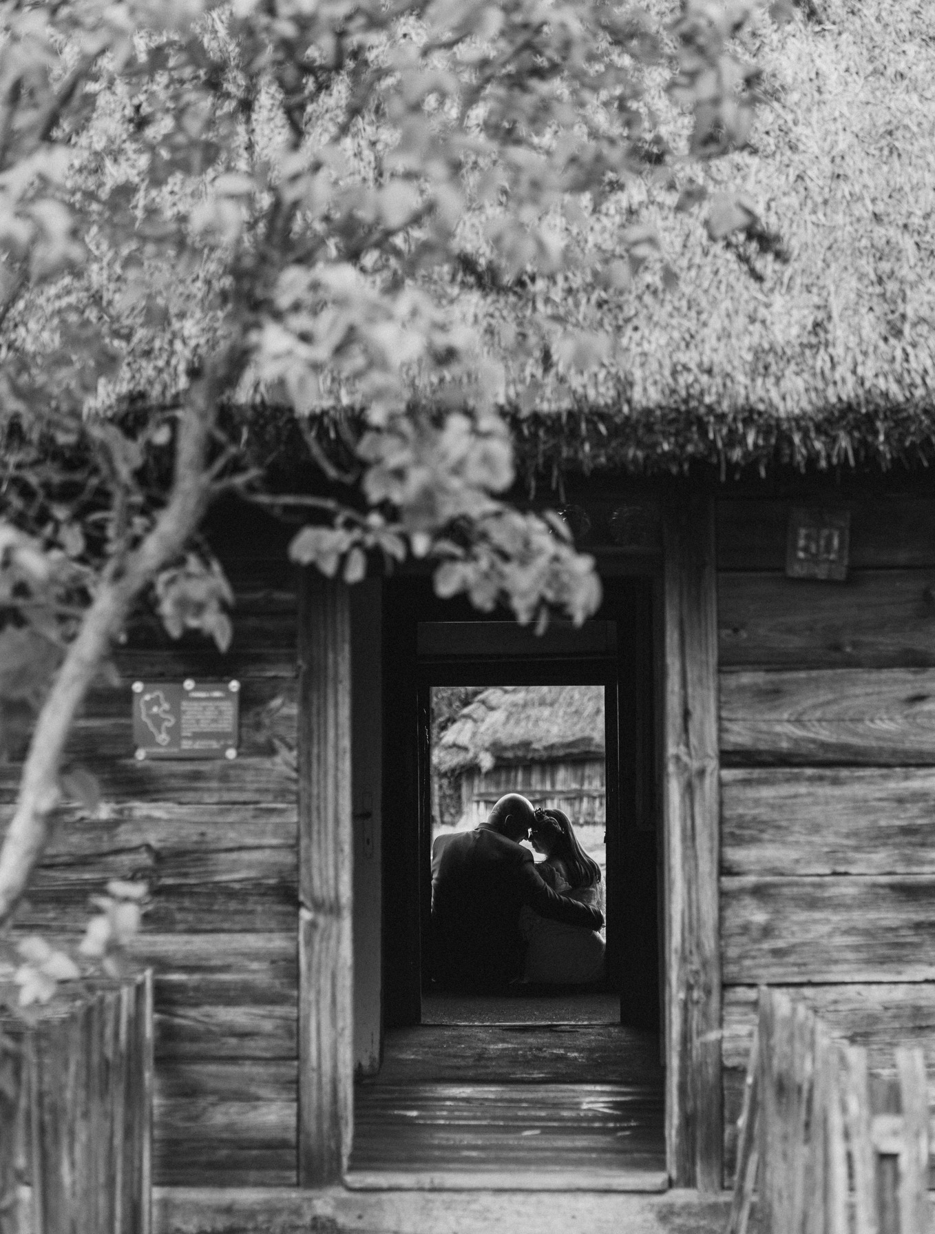 Czarno-białe zdjęcie pary w drzwiach drewnianej chaty z strzechą. Romantyczne ujęcie z bliska, z głębią ostrości na zakochanych.