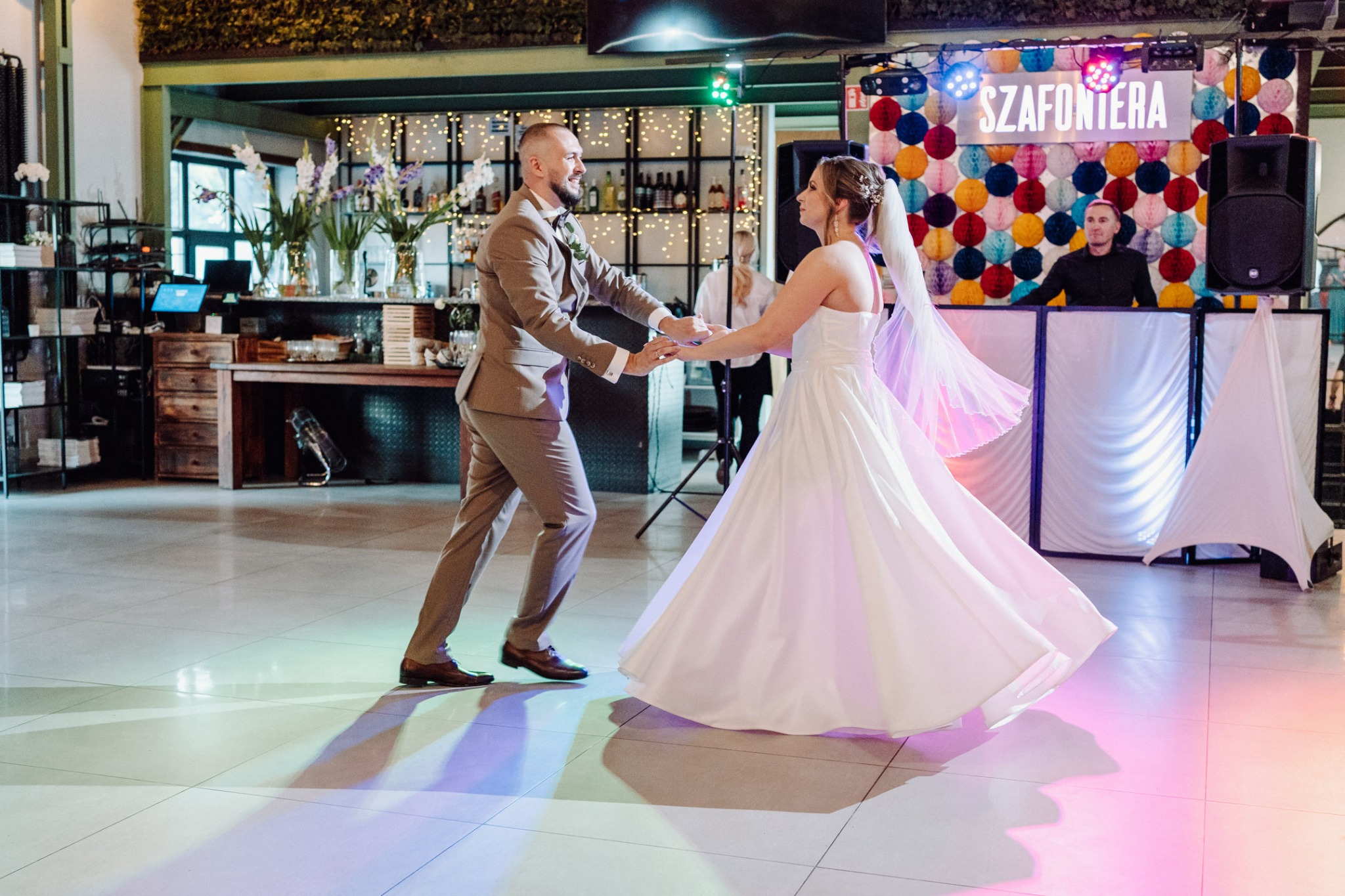 Uśmiechnięta para młoda tańczy na sali weselnej. W tle DJ z napisem 'Szafoniera' i dekoracje. Parkiet oświetlony kolorowymi światłami.