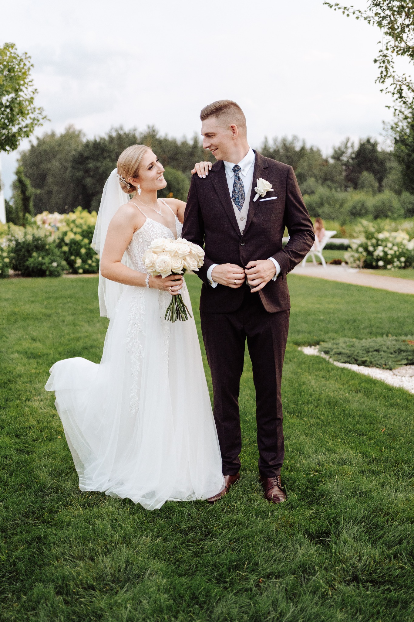 Para młoda: panna młoda w sukni z bukietem białych róż, pan młody w brązowym garniturze, spoglądają na siebie na tle zielonego ogrodu.