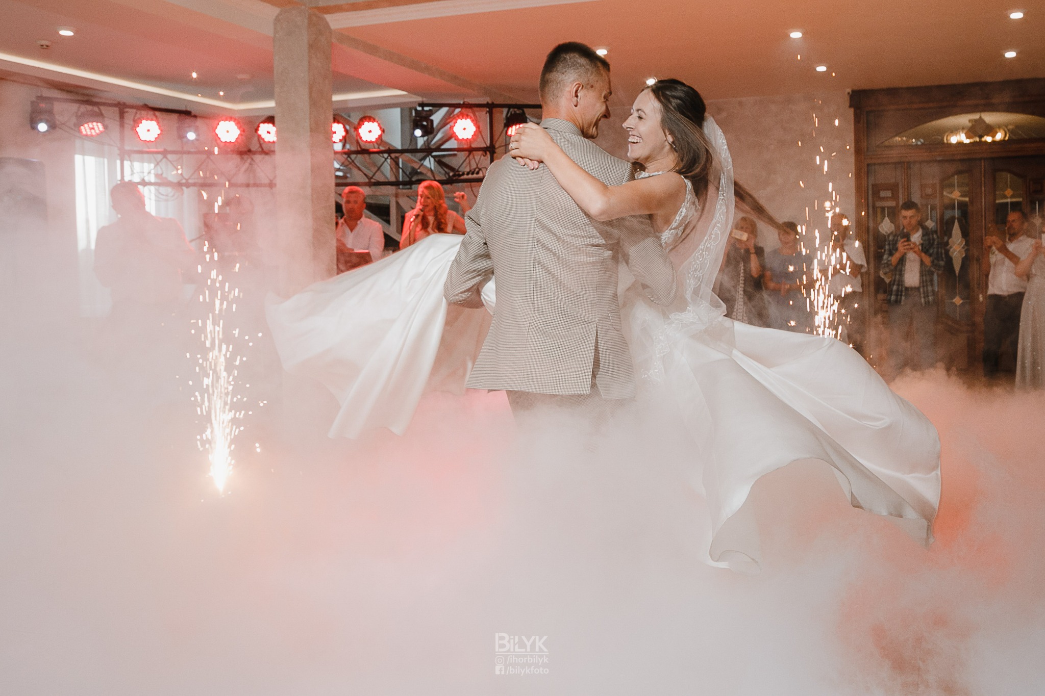 Para młoda tańczy w oparach dymu i blasku zimnych ogni podczas pierwszego tańca na sali weselnej, w tle goście.