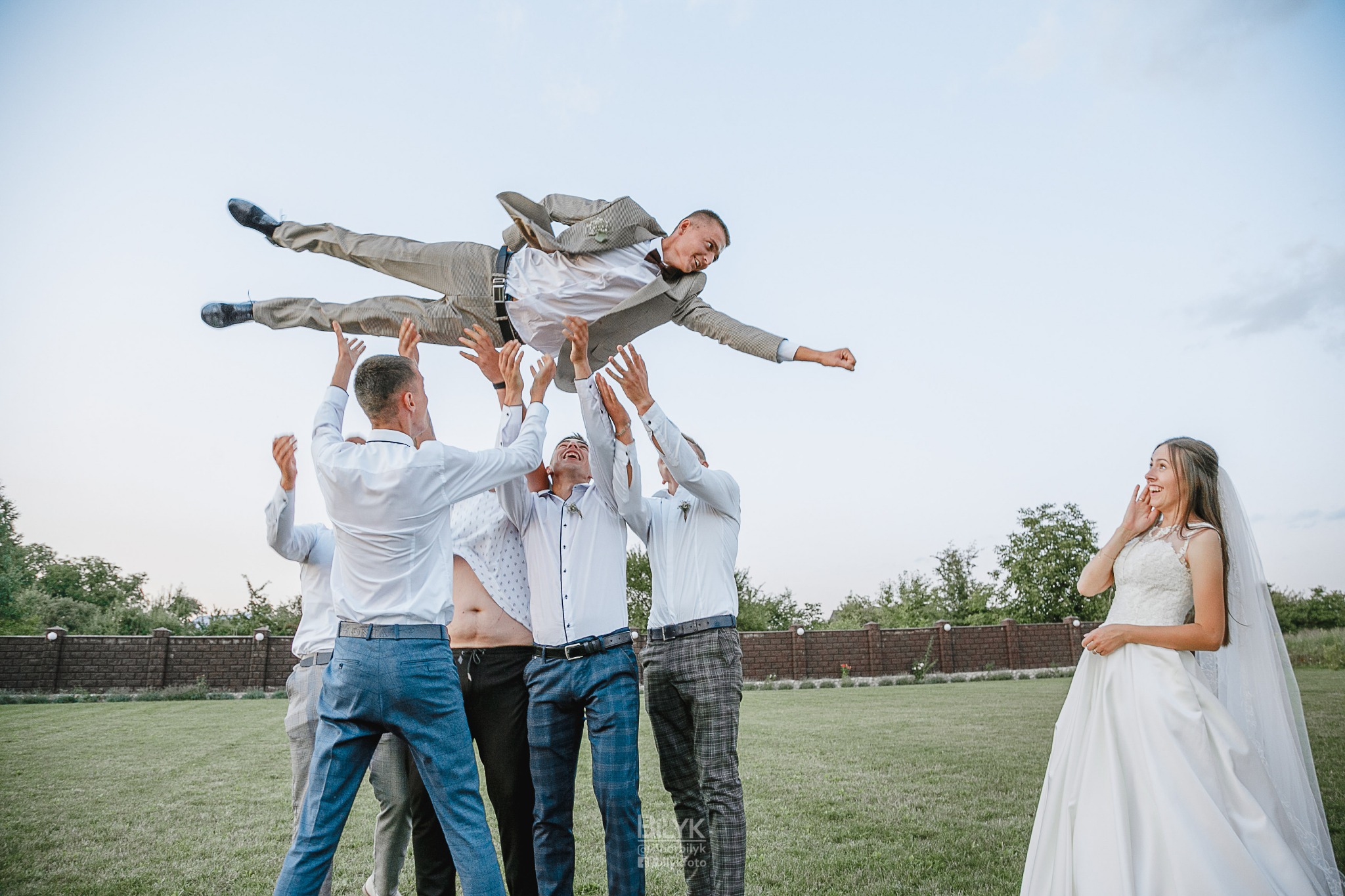 Pan młody w szarym garniturze, podrzucany do góry przez grupę mężczyzn w białych koszulach i spodniach, na tle uśmiechniętej panny młodej w białej sukni i welonie.