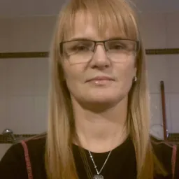 Portret kobiety w okularach z blond prostymi włosami i srebrnym naszyjnikiem w kształcie serca, na tle jasnych płytek ceramicznych.