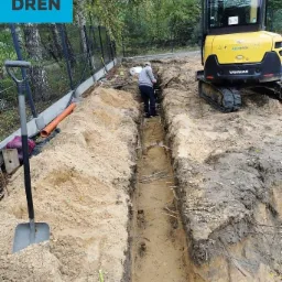 Wykopany rów w piaszczystej ziemi, obok żółta mini koparka Yanmar, widoczna osoba w rowie i szpadel wbity w ziemię, pomarańczowa rura na brzegu wykopu, teren budowy otoczony siatką.