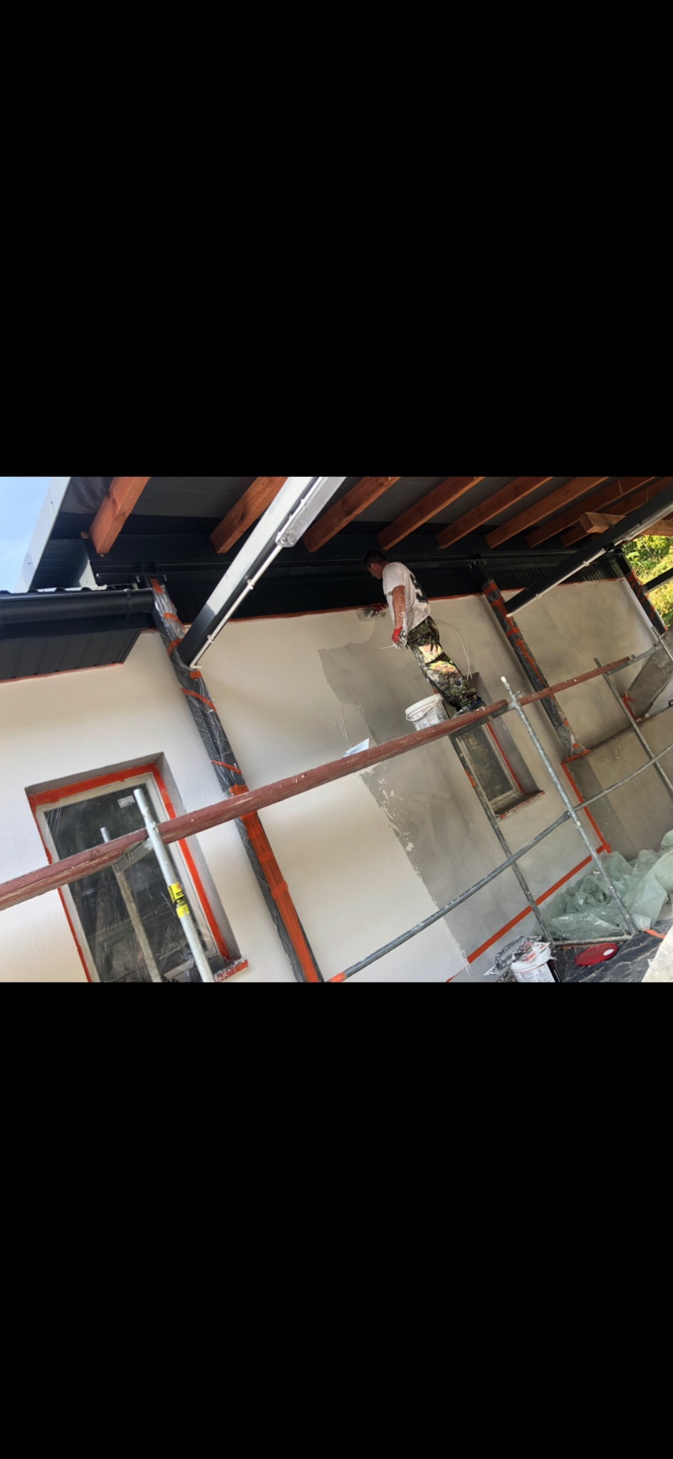 Pracownik na rusztowaniu, w trakcie nakładania szarego tynku na elewację budynku z oknami zabezpieczonymi folią, widoczne drewniane elementy konstrukcyjne dachu.