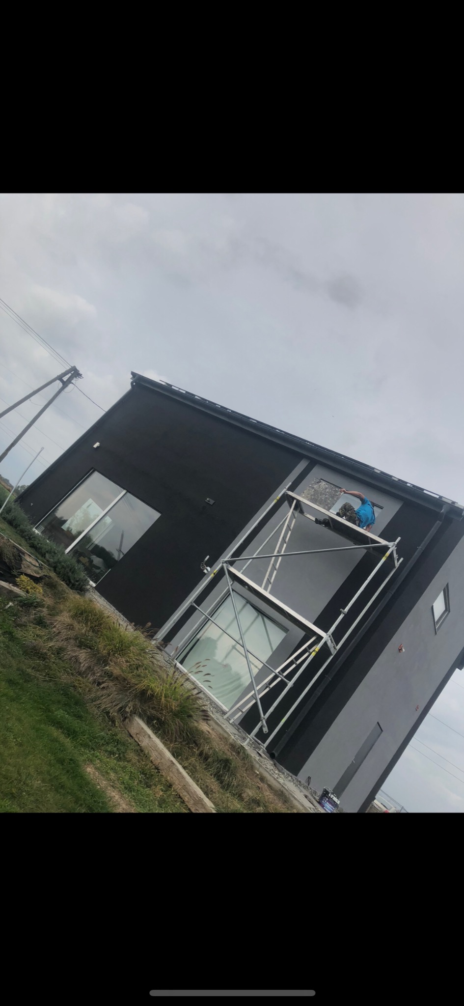 Pracownik na rusztowaniu przy czarnej, nowoczesnej elewacji domu jednorodzinnego, przygotowuje ścianę do dalszych prac wykończeniowych, widoczny fragment okna.