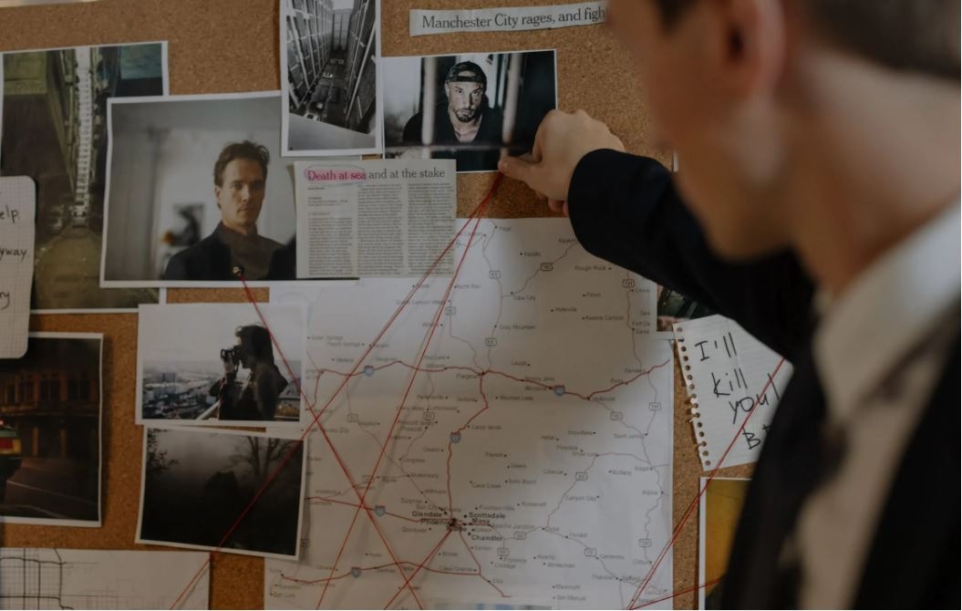 Tablica korkowa z mapą, zdjęciami i notatkami, połączona czerwonymi nitkami; mężczyzna w garniturze przypina zdjęcie, sugerując analizę sprawy.