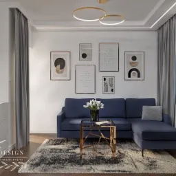 Salon z granatową sofą narożną, szarym dywanem, złotym stolikiem kawowym i galerią plakatów w ramkach na białej ścianie, oświetlony okrągłymi lampami sufitowymi i światłem dziennym.