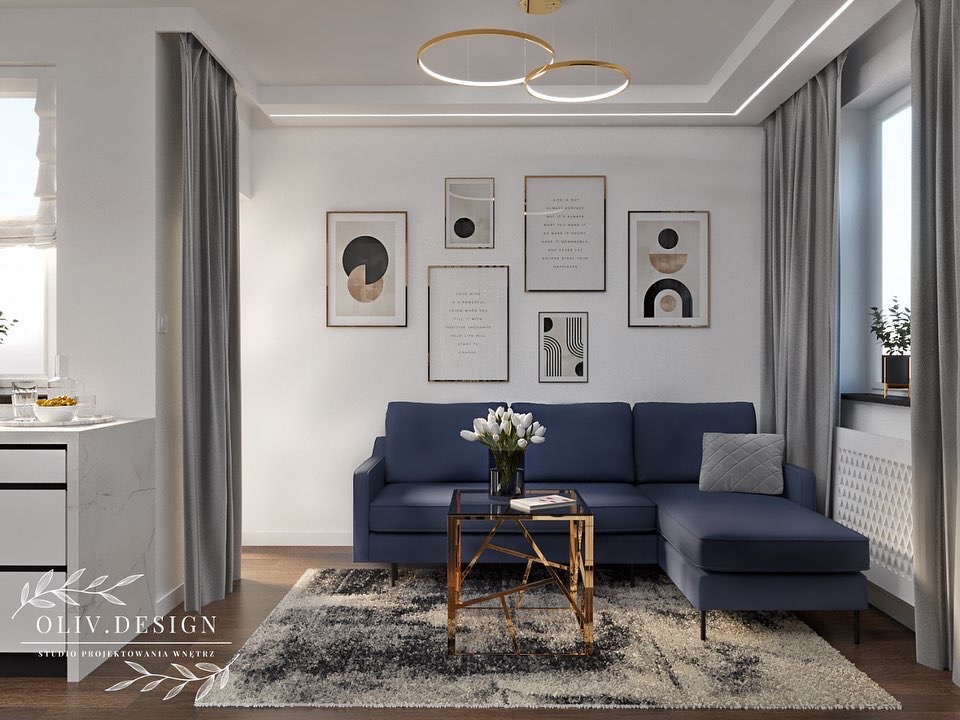 Salon z granatową sofą narożną, szarym dywanem, złotym stolikiem kawowym i galerią plakatów w ramkach na białej ścianie, oświetlony okrągłymi lampami sufitowymi i światłem dziennym.