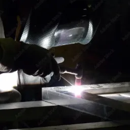 Spawacz w masce ochronnej łączy metalowe elementy, widoczny jasny łuk elektryczny i iskry.