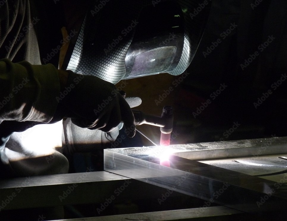 Spawacz w masce ochronnej łączy metalowe elementy, widoczny jasny łuk elektryczny i iskry.