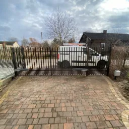 Czarna, metalowa brama przesuwna z automatyką, częściowo otwarta, z widocznym napędem i centralą sterującą. W tle biały samochód dostawczy z drabiną na dachu, zaparkowany na posesji z domem...