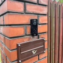 Czarny wideodomofon Eura zamontowany na ceglanym słupku obok brązowej skrzynki na listy z symbolem poczty, widoczny fragment drewnianego ogrodzenia.