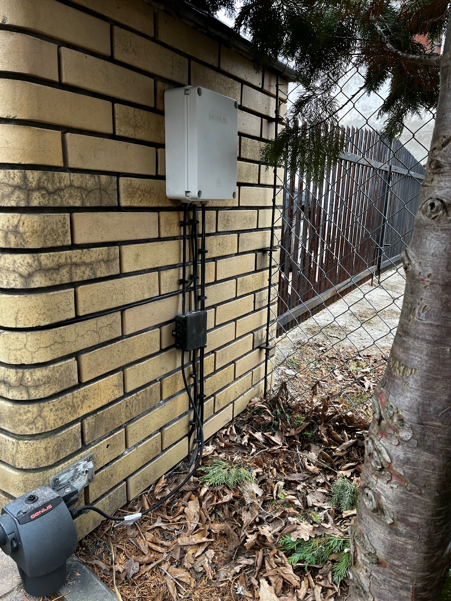 Zamontowany automat do bramy dwuskrzydłowej Genius G-Bat 300 firmy FAAC, z widoczną skrzynką sterującą na ceglanym słupku, okablowaniem w peszlach i opadłymi liśćmi u podstawy.