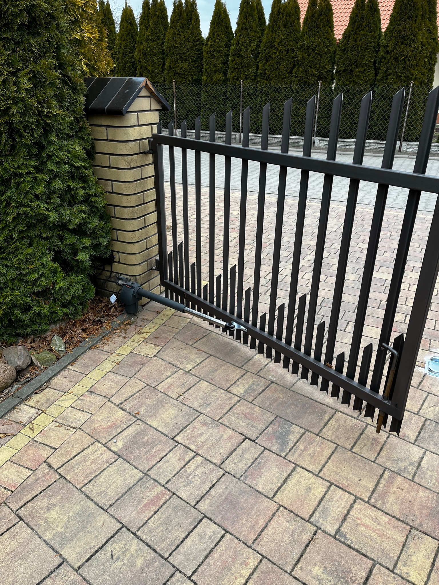 Ciemna, metalowa brama dwuskrzydłowa z zamontowanym siłownikiem FAAC Genius G-Bat 300, widoczna na tle brukowanej nawierzchni i kolumny z cegły klinkierowej, otoczona zielenią.