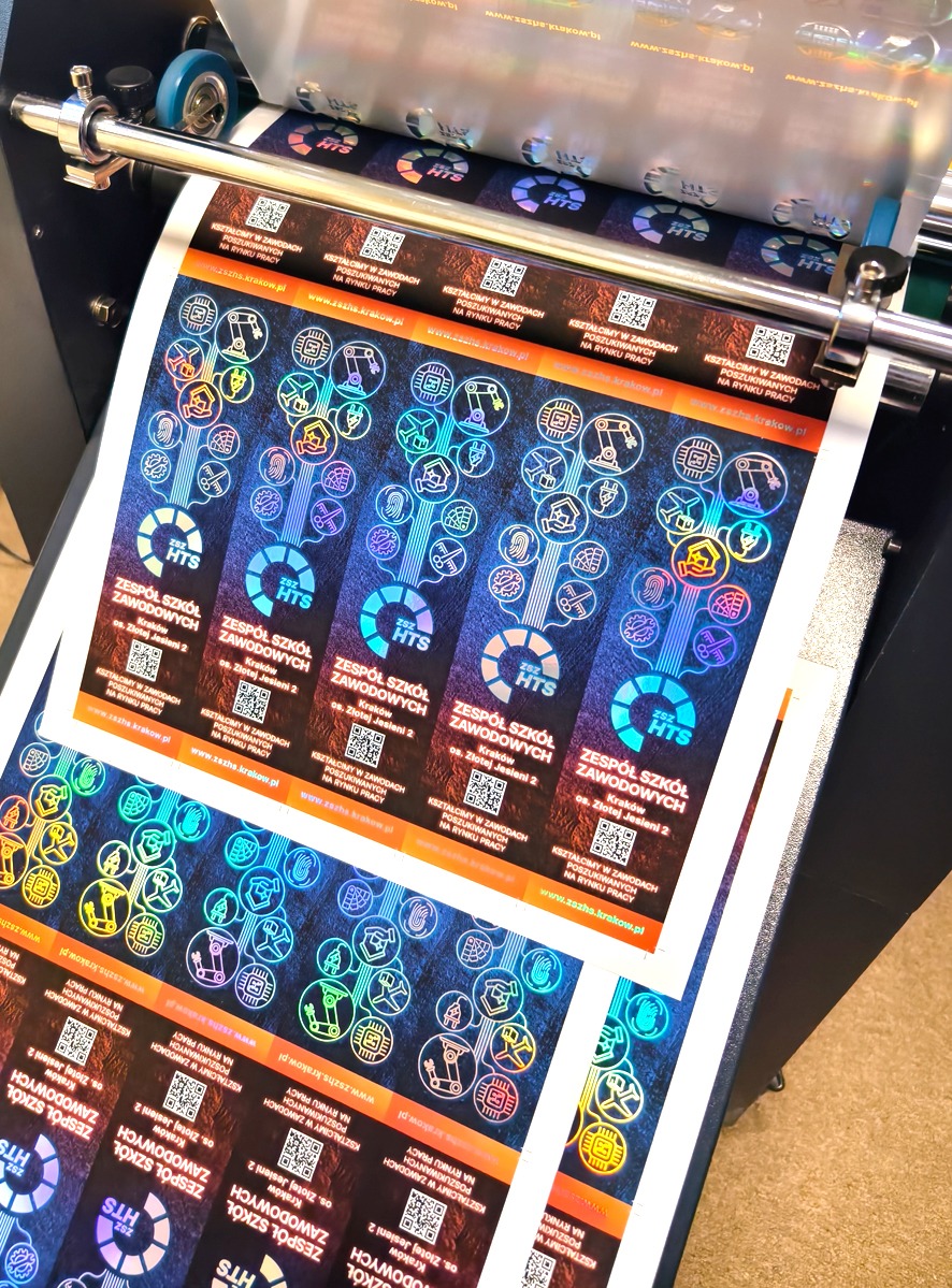 Arkusz zadrukowanych zakładek do książek z folią holo, wychodzący z maszyny drukarskiej. Widoczne elementy graficzne i tekstowe, w tym logo szkoły zawodowej.