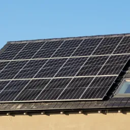 Panele słoneczne zamontowane na dachu domu z dwoma oknami dachowymi, widoczne z dołu na tle błękitnego nieba.