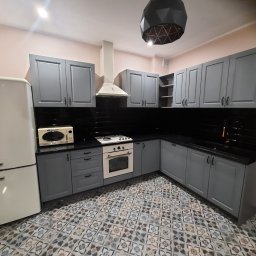 TomMeb Tomasz Kucharski - Kompletna kuchnia w szarych szafkach z czarnym blatem i beżowym sprzętem AGD na wzorzystej podłodze.