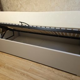 TomMeb Tomasz Kucharski - Otwarta szafa wnękowa z mechanizmem podnoszenia materaca, ukazująca przestrzeń do przechowywania. Widoczny materac i drewniana podłoga.