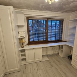 TomMeb Tomasz Kucharski - Biała zabudowa meblowa z szafami, szafkami i półkami, wkomponowana w przestrzeń okienną z drewnianymi żaluzjami. Klasyczny styl, jasne wnętrze.
