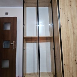 TomMeb Tomasz Kucharski - Wysoka szafa z przeszklonymi drzwiami w czarnej ramie, z półkami w kolorze drewna, na kółkach. Obok szafa z drewnianymi frontami i czarnymi obramowaniami.