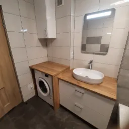 Nowoczesna łazienka z zabudową pralki i umywalki na drewnianym blacie, białe szafki i płytki, lustro z oświetleniem.