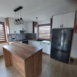 Nowoczesna kuchnia z wyspą, białymi frontami szafek, drewnianymi akcentami i geometrycznymi lampami wiszącymi.