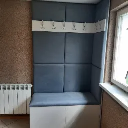 Narożna zabudowa tapicerowana w przedpokoju z siedziskiem i wieszakami na ścianie, jasnoszara kolorystyka, białe szafki pod siedziskiem, grzejnik obok.