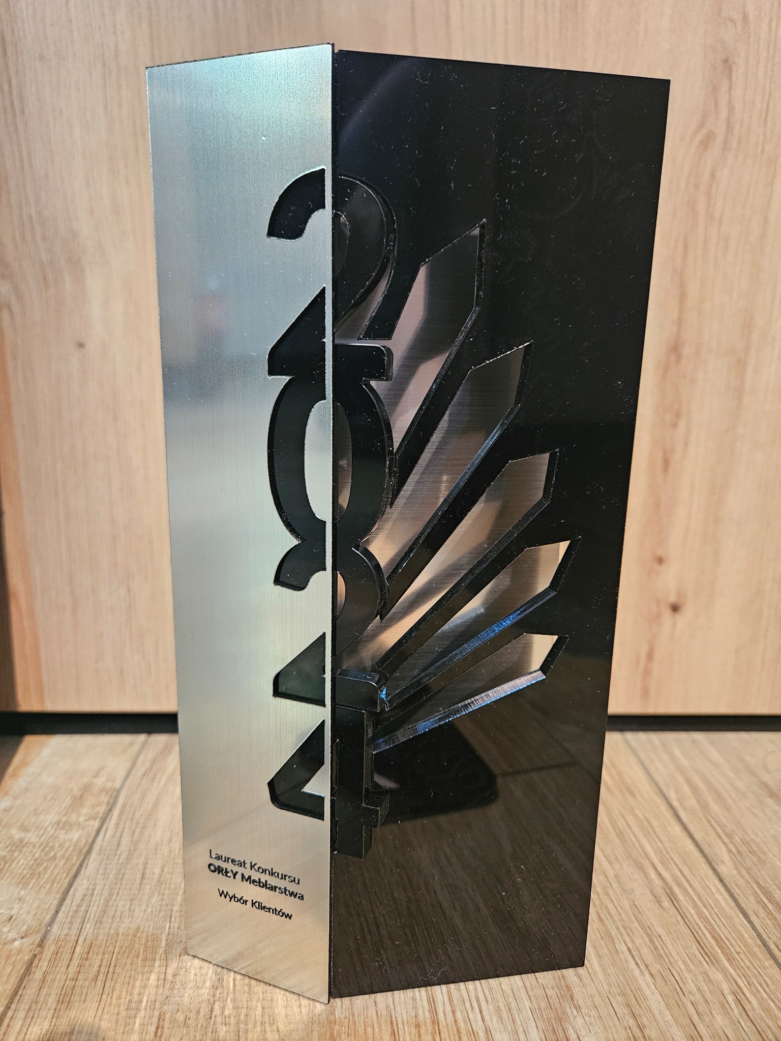 Statuetka konkursu 'Orły Meblarstwa - Wybór Klientów' z wyciętymi numerami '2024' i symbolem skrzydeł, srebrno-czarna, na drewnianym podłożu.