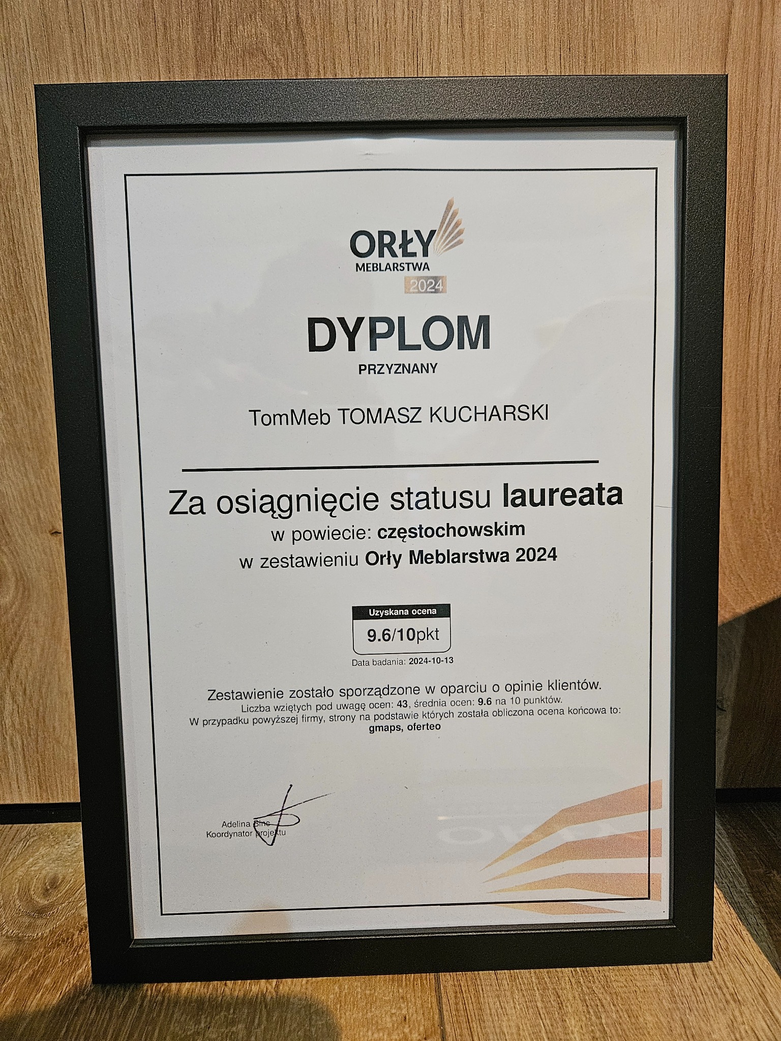 Dyplom Orły Meblarstwa 2024 przyznany firmie TomMeb Tomasz Kucharski za osiągnięcie statusu laureata w powiecie częstochowskim, ocena 9.6/10 pkt, czarna ramka na drewnianym tle.