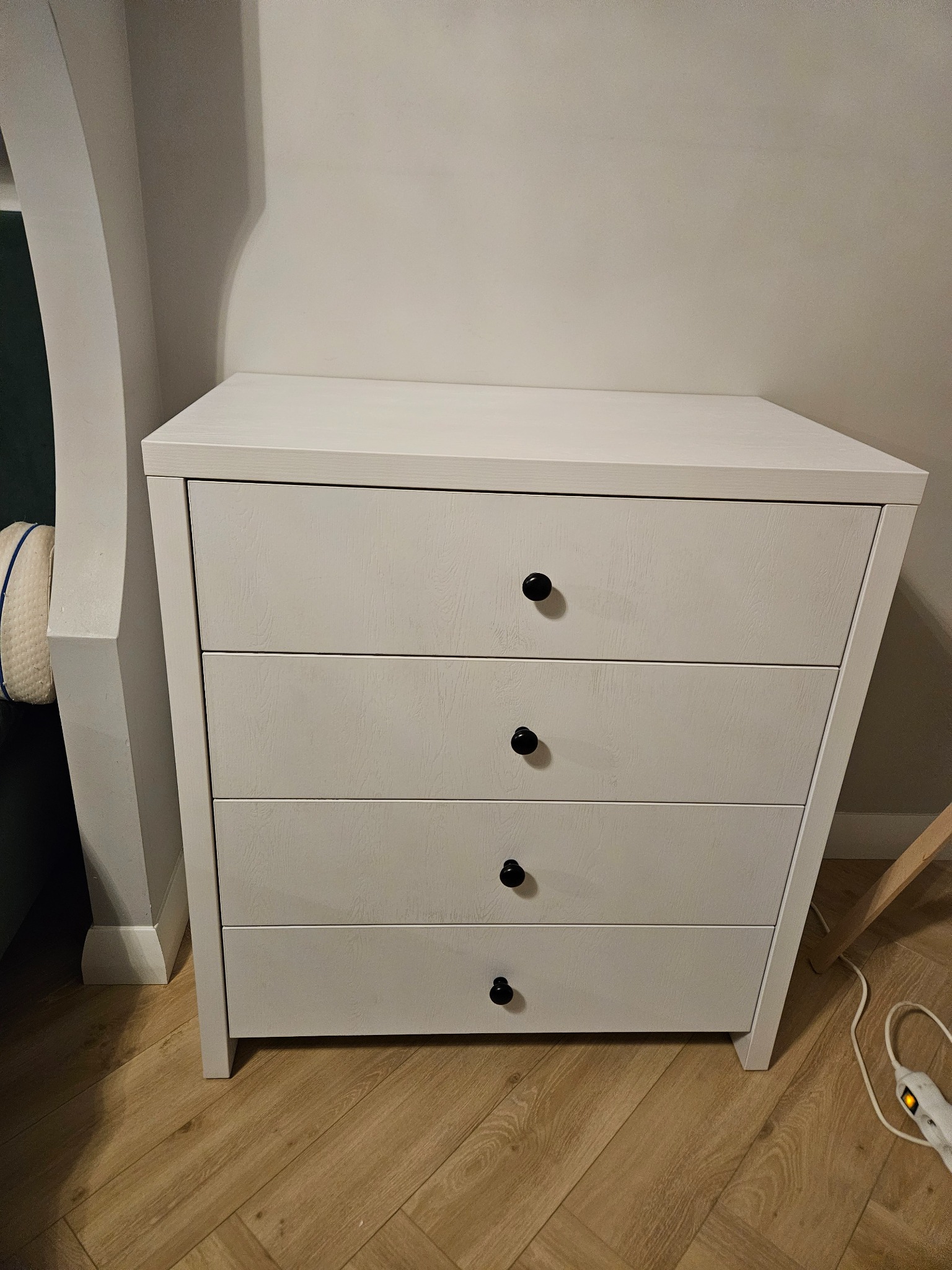 Biała komoda z czterema szufladami i okrągłymi, czarnymi uchwytami, stojąca na drewnianej podłodze w rogu pokoju. Widoczny fragment sofy i listwy przypodłogowej.