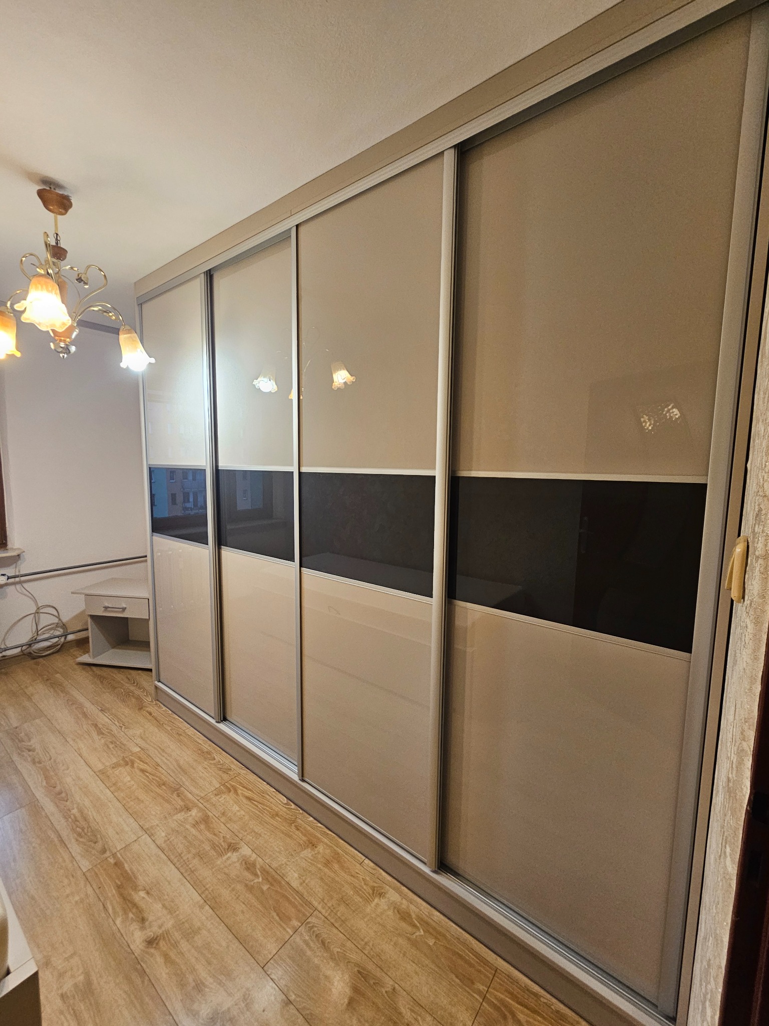 Beżowa szafa wnękowa z przesuwnymi drzwiami, z poziomym pasem w kolorze czarnym, na drewnianej podłodze. Widoczny fragment żyrandola i stolika.