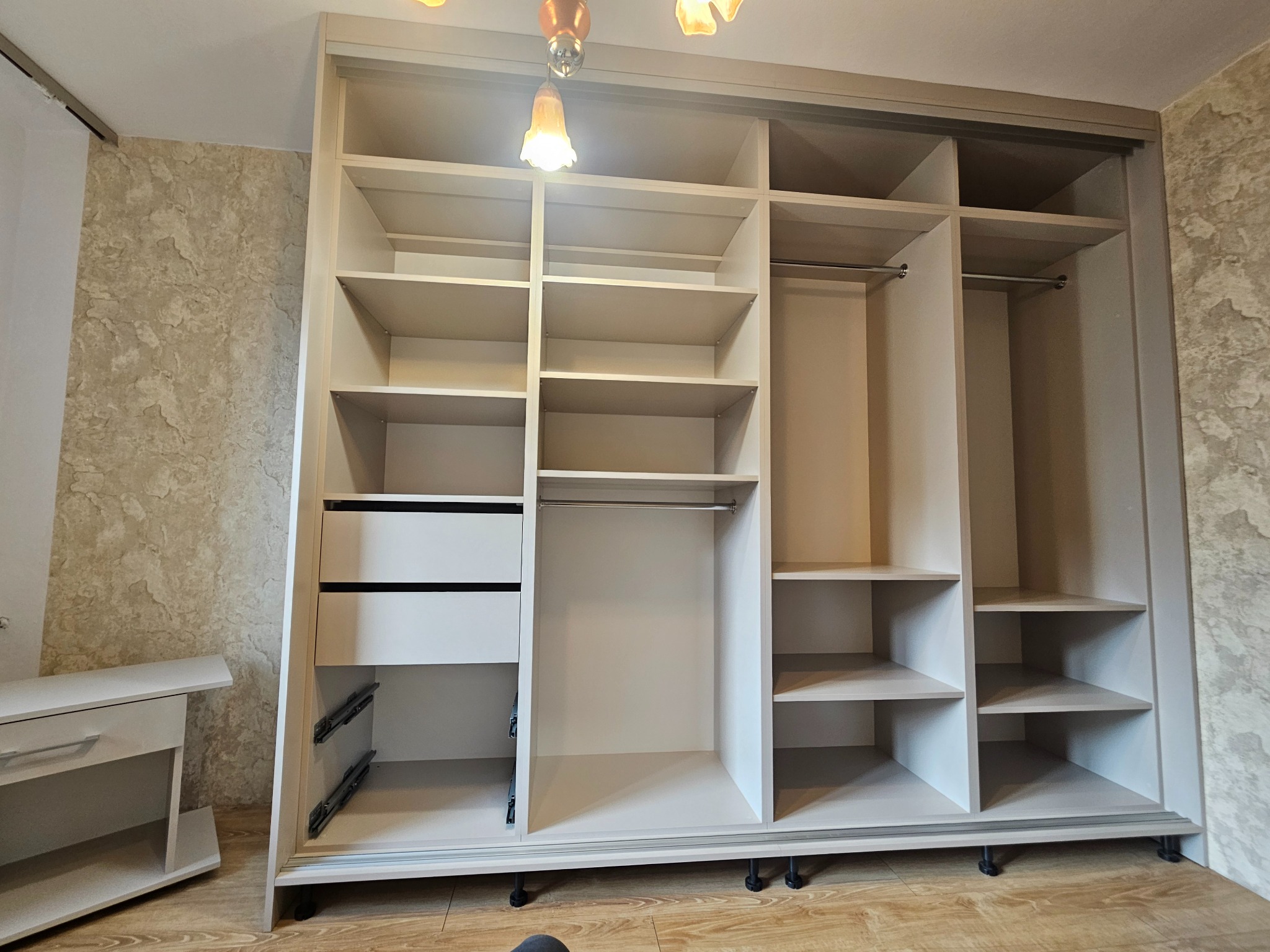 Jasna, otwarta szafa wnękowa z przesuwnymi drzwiami, widoczne półki, szuflady i drążki na ubrania. Ustawiona na drewnianej podłodze, przy ścianie o wzorzystej tapecie.