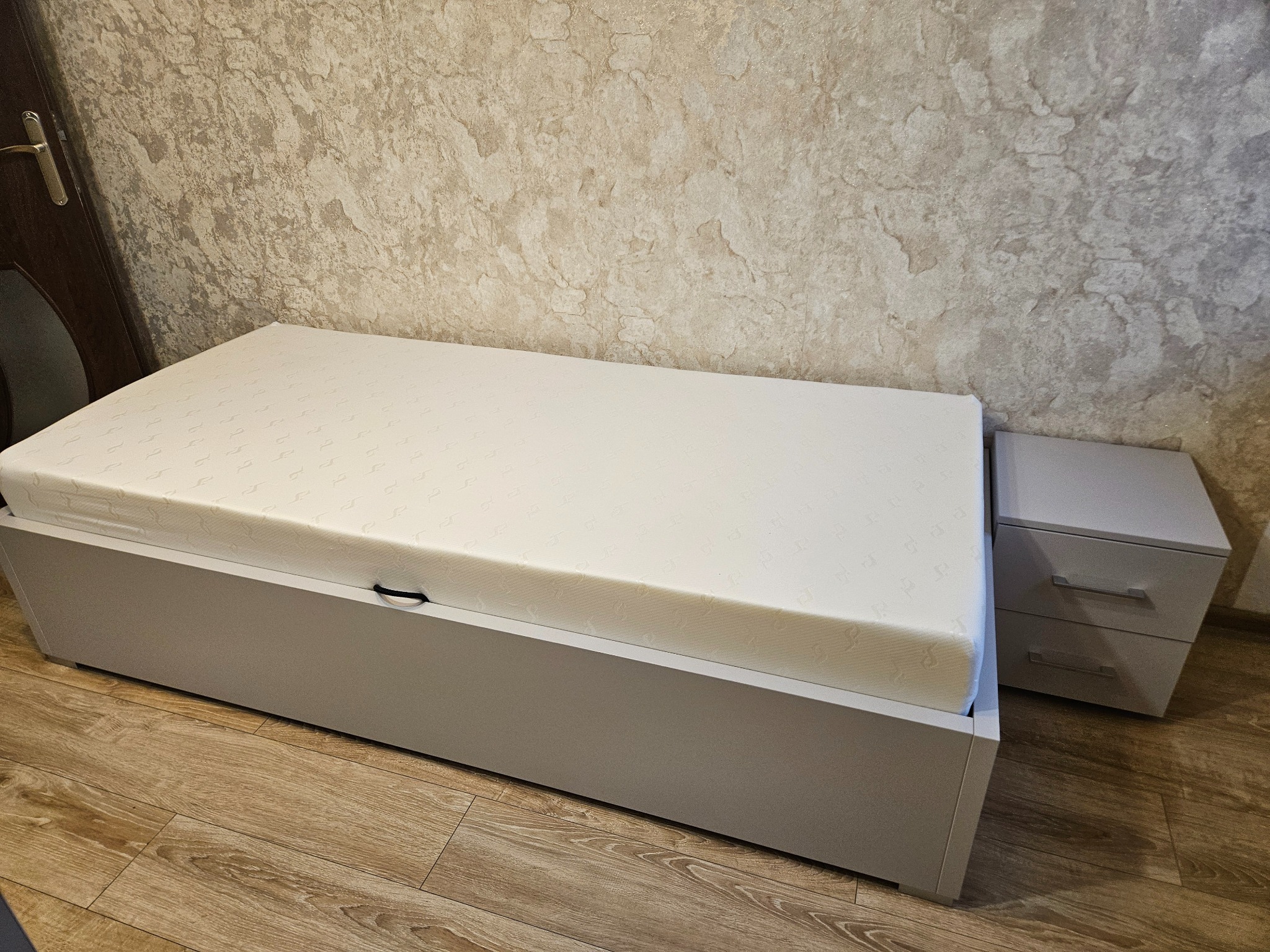 Szare łóżko z pojemnikiem i materacem, obok szafka nocna z dwiema szufladami. Całość w neutralnej kolorystyce, na drewnianej podłodze.