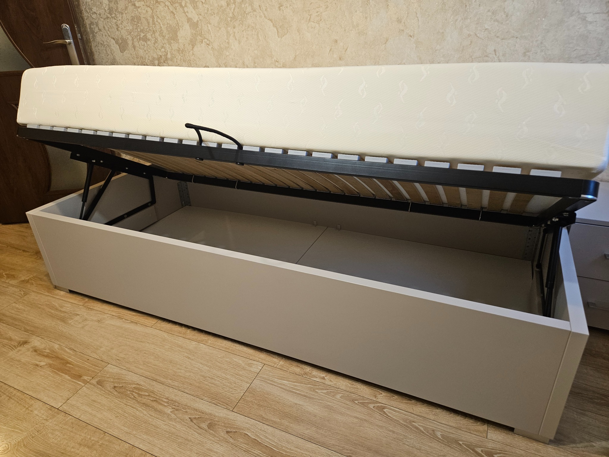 Otwarta szafa wnękowa z mechanizmem podnoszenia materaca, ukazująca przestrzeń do przechowywania. Widoczny materac i drewniana podłoga.