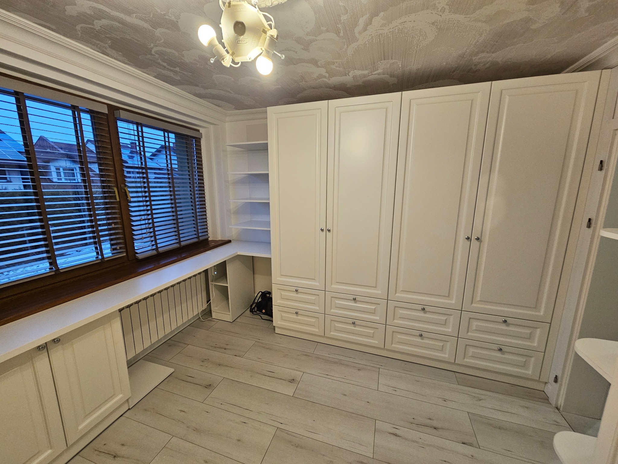 Biała szafa wnękowa z szufladami i półkami, wbudowana w pokoju z oknem i drewnianymi żaluzjami. Widoczny fragment biurka i grzejnika pod oknem.