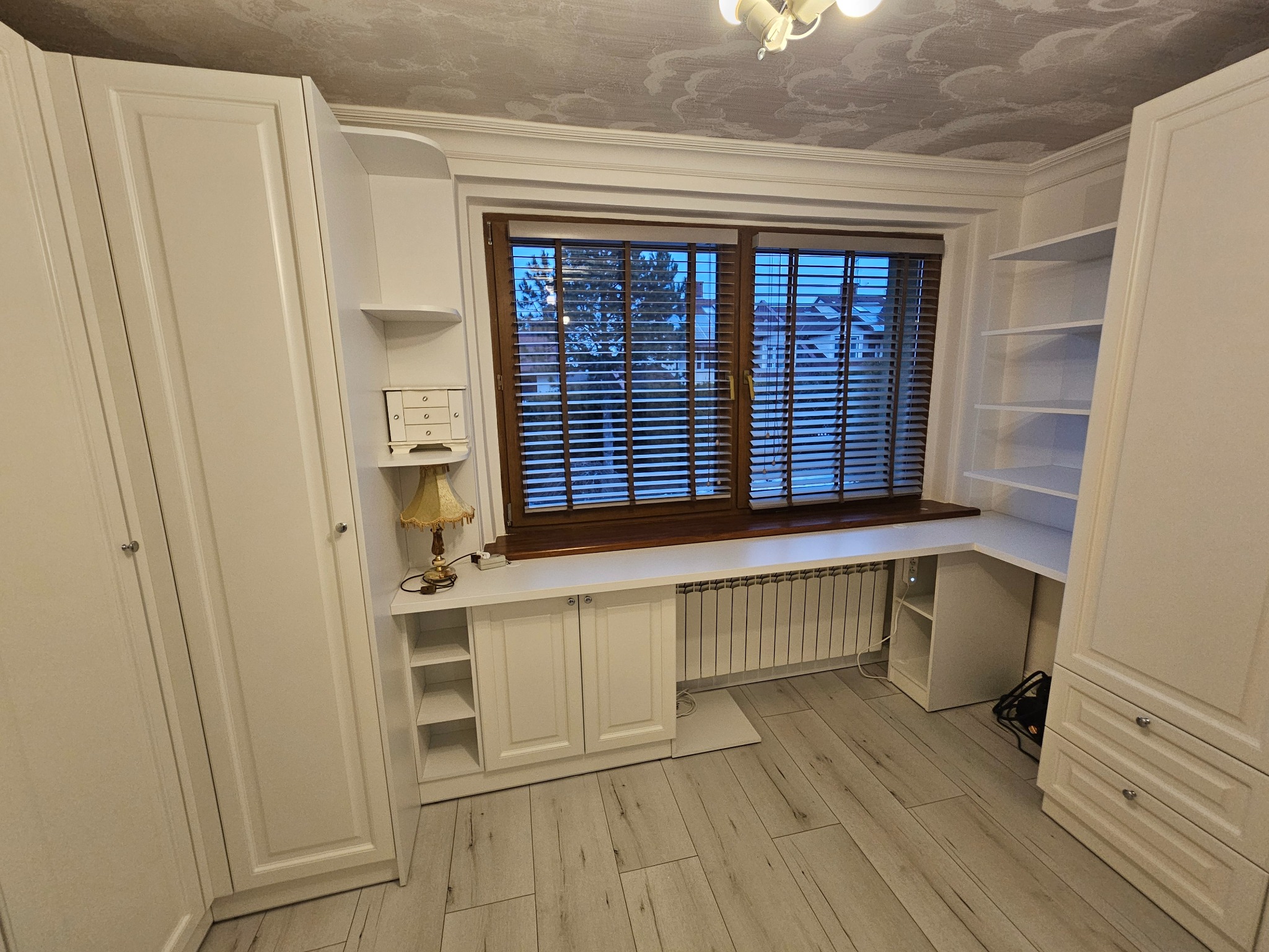 Biała zabudowa meblowa z szafami, szafkami i półkami, wkomponowana w przestrzeń okienną z drewnianymi żaluzjami. Klasyczny styl, jasne wnętrze.