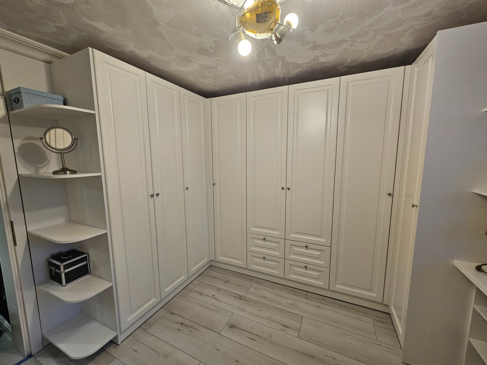 Biała szafa wnękowa z półkami, oświetlona lampą sufitową. Widoczne detale wykończenia i drewnopodobna podłoga. Klasyczny design.