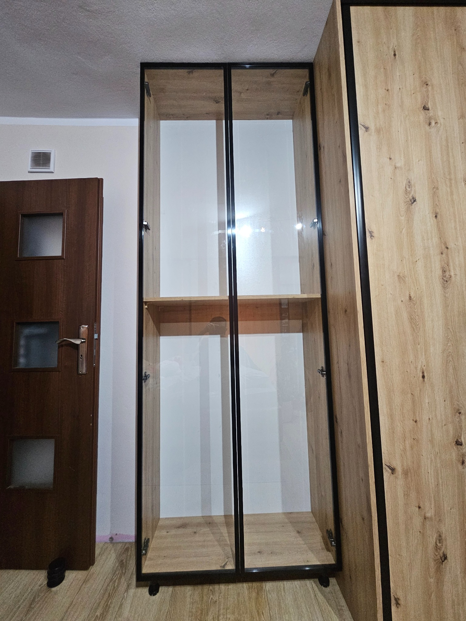 Wysoka szafa z przeszklonymi drzwiami w czarnej ramie, z półkami w kolorze drewna, na kółkach. Obok szafa z drewnianymi frontami i czarnymi obramowaniami.