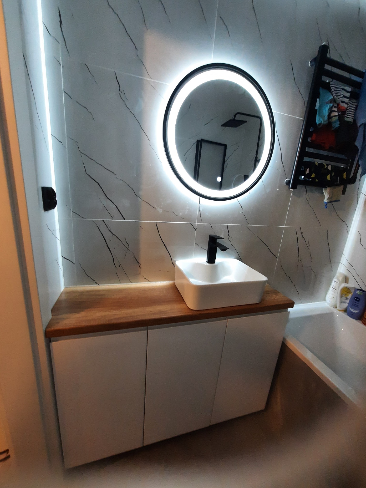 Nowoczesna łazienka z szafką pod umywalką z drewnianym blatem, okrągłym lustrem z podświetleniem LED i marmurowymi płytkami na ścianie.