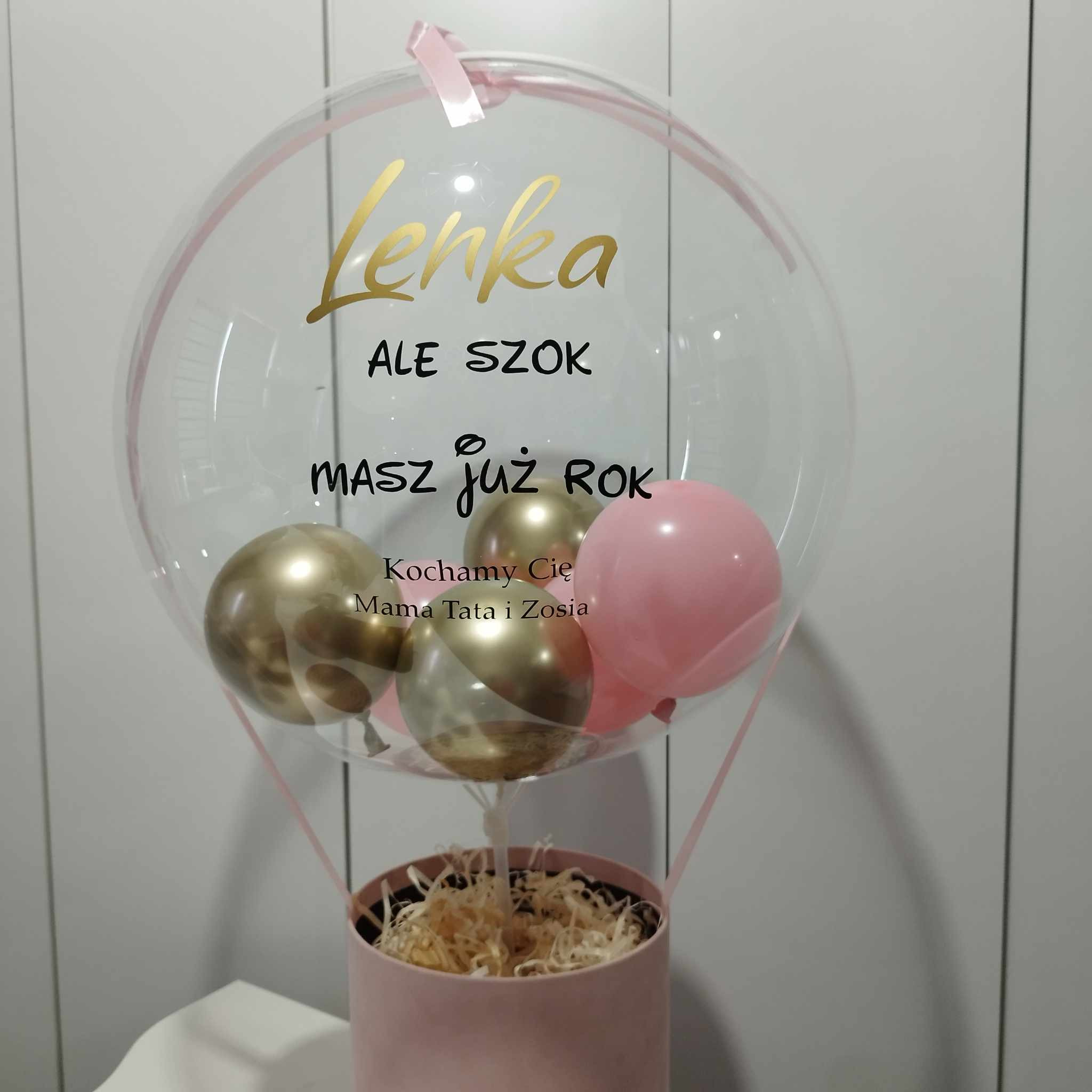 Przezroczysty balon z napisem 'Lenka ALE SZOK masz już ROK Kochamy Cię Mama Tata i Zosia', zawierający złote i różowe balony, umieszczony w różowym pudełku wypełnionym siankiem.