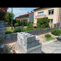 Częściowo ukończony murek z szarych betonowych bloczków, paleta z zapasem bloczków, widok na dom z ogrodem w tle.