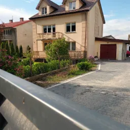 Dwupiętrowy dom z beżową elewacją, drewnianymi balkonami i garażem z czerwonymi wrotami, widoczny zza metalowego ogrodzenia, z zadbanym ogrodem i kostką brukową na podjeździe.