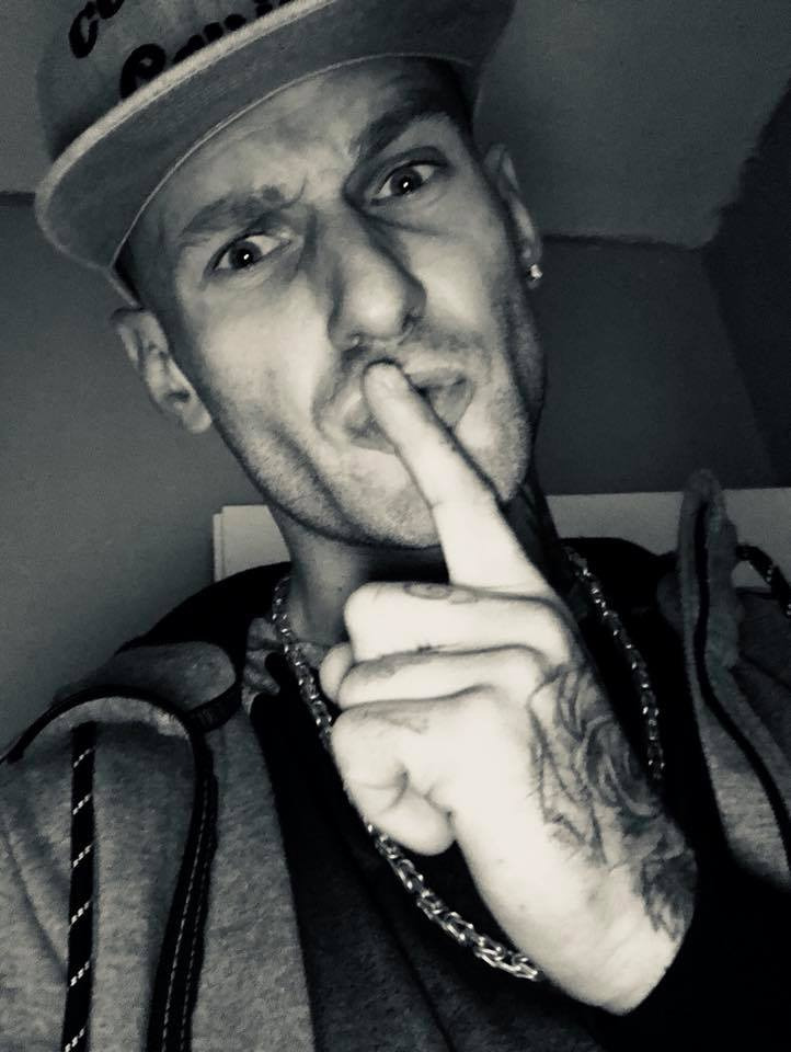 Czarno-białe selfie mężczyzny w czapce z daszkiem i łańcuchem, z tatuażem na dłoni, przykładającego palec do ust w geście uciszenia.