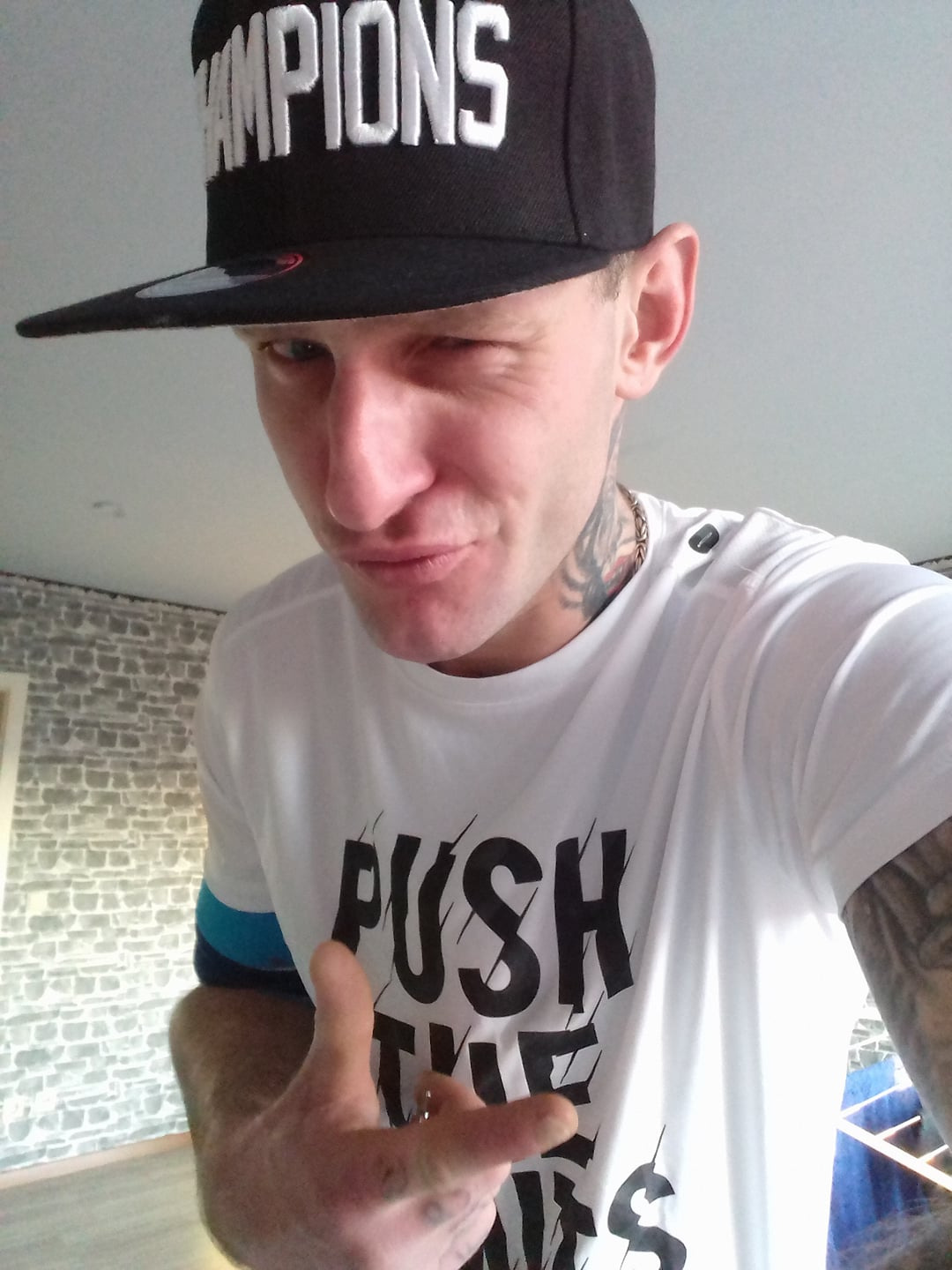 Mężczyzna w czapce z napisem 'Champions' i białej koszulce z napisem 'Push the limits', mruga okiem, z tatuażami na szyi i ramieniu, wnętrze z imitacją ściany z cegieł w tle.