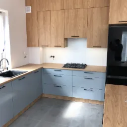 Jasna kuchnia z drewnianymi szafkami górnymi i błękitnymi dolnymi, czarny zlew i płyta gazowa, widoczne niedokończone gniazdka elektryczne.