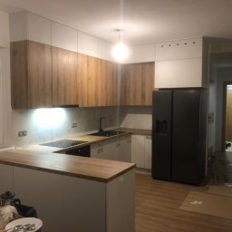 Stanislav Alimgulov - Nowoczesna kuchnia z drewnianymi frontami szafek górnych i białymi dolnymi, czarnym zlewem i płytą indukcyjną, oświetleniem podszafkowym, czarną lodówką side-by-side, częściowo rozpakowana...