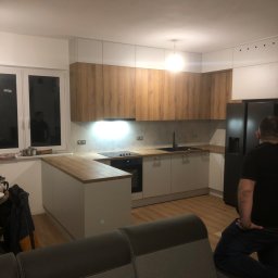 Stanislav Alimgulov - Nowoczesna kuchnia z drewnianymi szafkami górnymi i białymi dolnymi, wyspa z drewnianym blatem, czarna lodówka side-by-side, widok z salonu na częściowo umeblowaną przestrzeń.