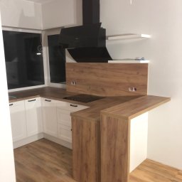 Stanislav Alimgulov - Nowoczesna kuchnia z białymi szafkami i drewnianymi blatami, czarny okap, drewniana podłoga, widoczne gniazdka elektryczne w panelu nad blatem.